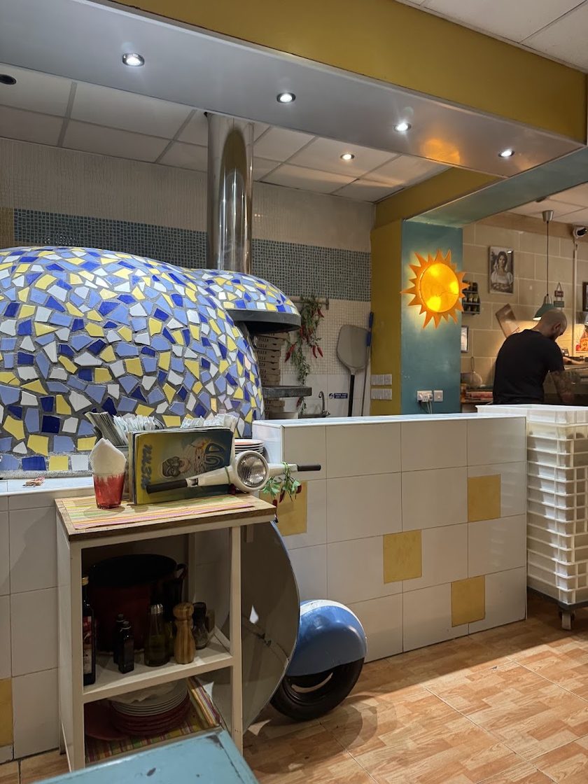 The wood oven's blue and yellow mosaic dome beside the service counter, with a metal sun on the wall, hanging dried chillies and the front half of a pale blue scooter built into the counter