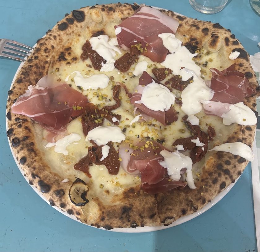 A white pizza with Parma ham, torn stracciatella, roast aubergine and pistachio crumb, seen from above on a turquoise table