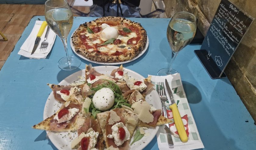 A turquoise table seen from above with a margherita pizza, a plate of pizza wedges, two wine glasses and a small chalkboard