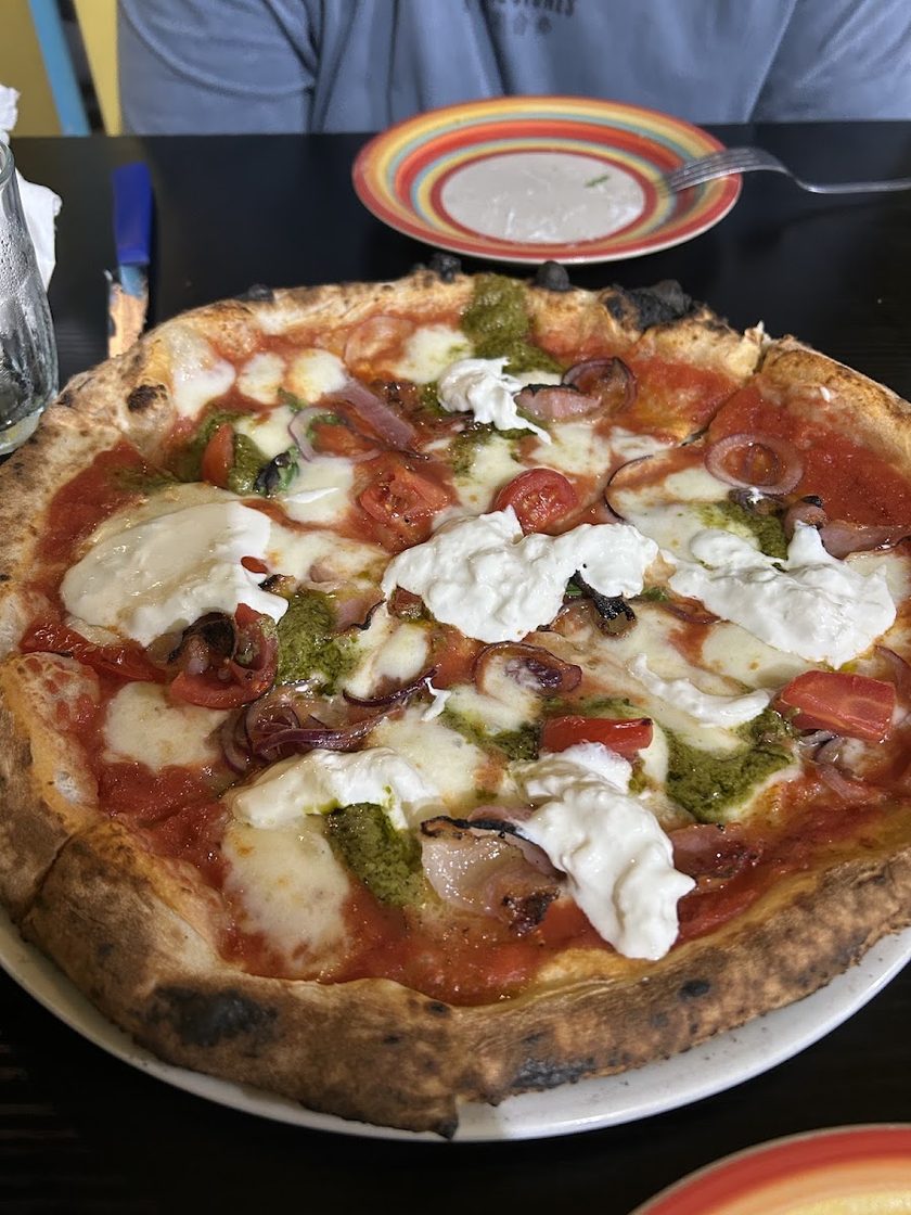 A pizza with torn stracciatella, dabs of pesto, cherry tomatoes and black olives, on a white plate on a dark table