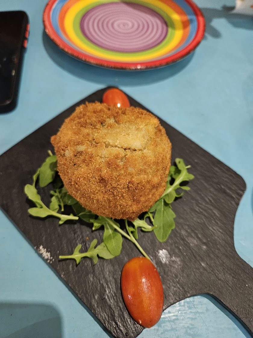 A large breaded and deep-fried ball on a slate board with rocket and a tomato, on a turquoise table beside a rainbow-striped plate