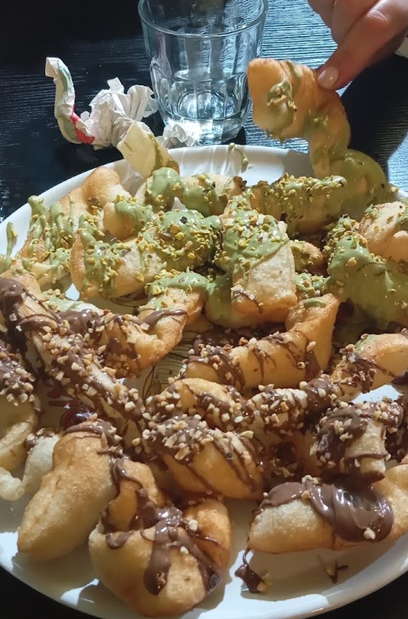 A plate of fried pastry pieces, some rolled in nuts and chocolate and some dusted with pistachio, with a hand reaching in