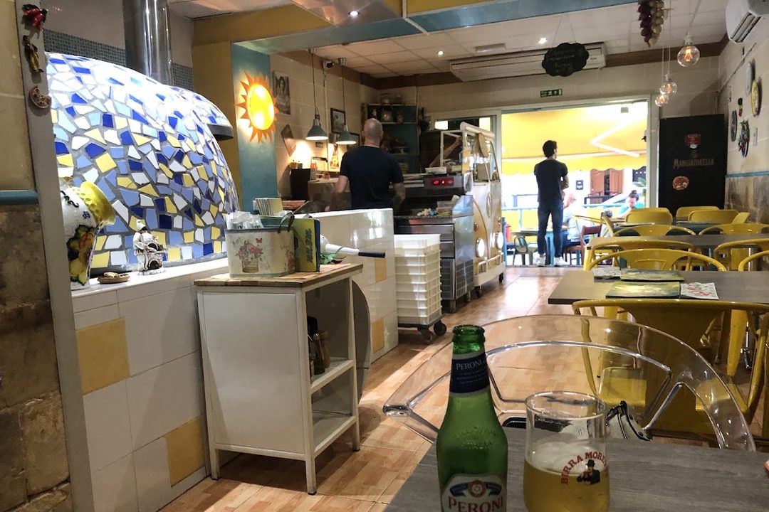 The dining room at Made in Sud, with the mosaic tiled wood oven on the left, the pizzaiolo at the counter and yellow chairs at grey tables beyond
