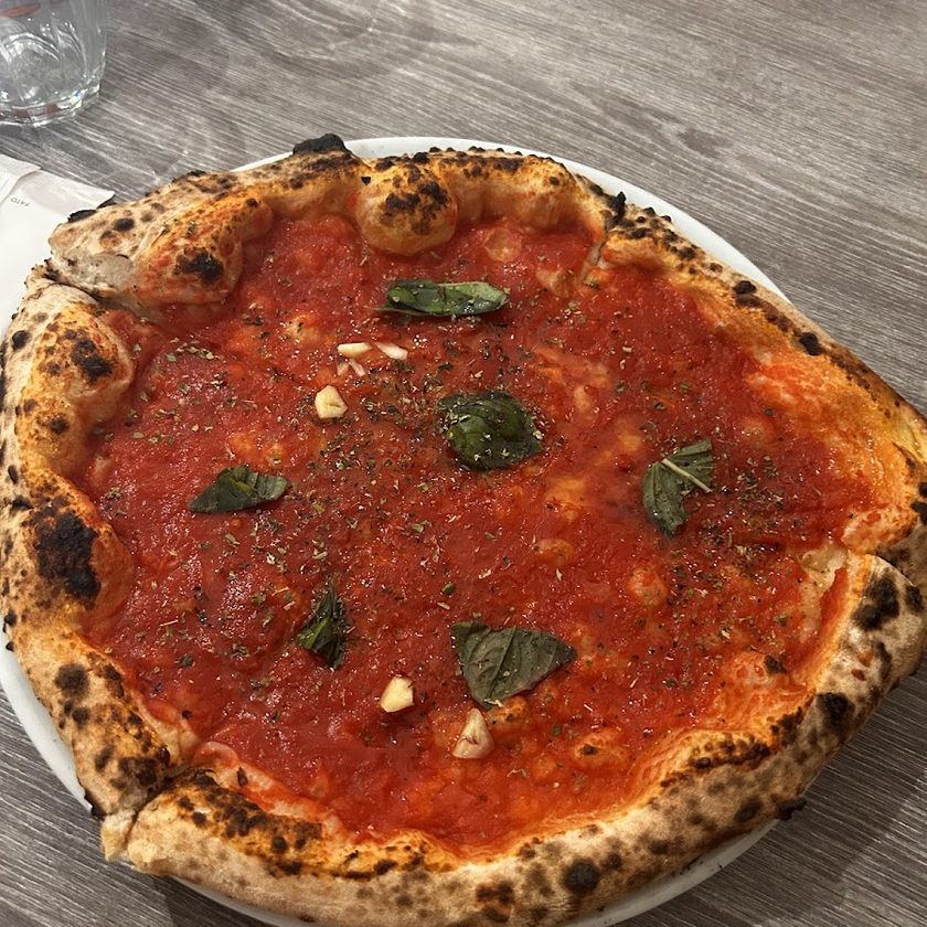 A marinara pizza with no cheese, tomato, garlic, oregano and basil on a blistered leopard-spotted crust, on a white plate on a grey wood table