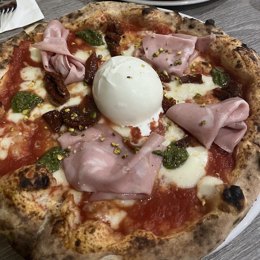A pizza with folded mortadella, a whole burrata, pistachio crumb and sun-dried tomatoes, with a second pizza on the table behind it