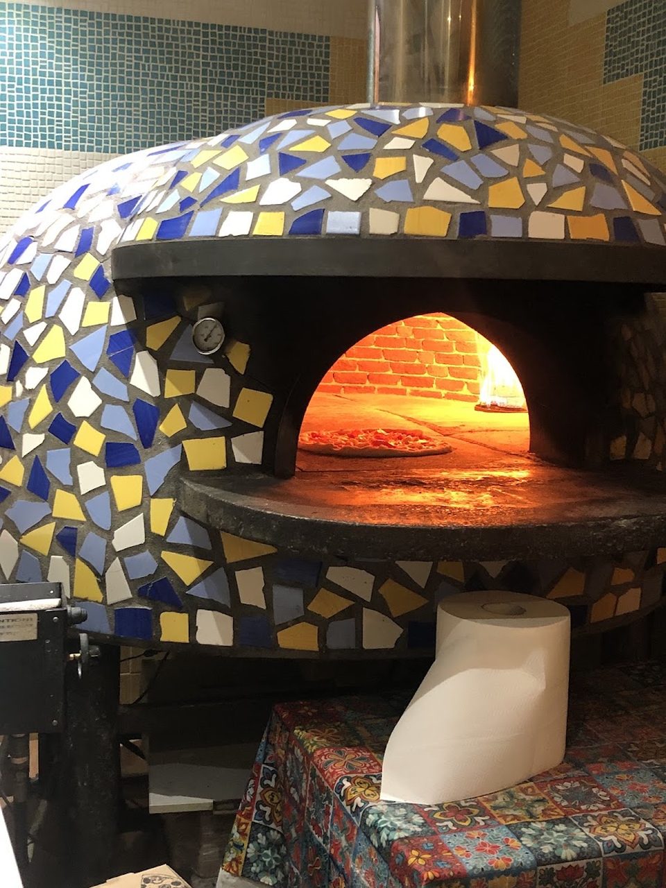 The wood oven's dome, covered in broken cobalt, yellow and white mosaic tile, with a pizza cooking on the floor of the fire-lit brick chamber inside